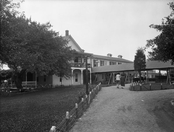 View up driveway toward a house and covered walkway. A fence is along the road, and two women walking up to the house, where a group of women are sitting on the porch.