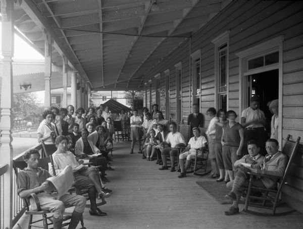People posing on the porch. Some of the people are standing, and others are sitting in rocking chairs. Part of the covered walkway can be seen in the left background.