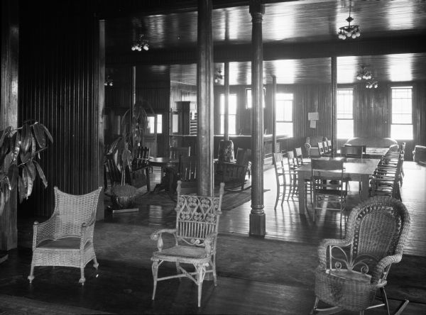 Interior view of the lobby, with three wicker chairs sitting in a row in the foreground, and a large room behind it that includes the front desk and numerous tables and chairs. Carved wooden beams, electric light fixtures, wooden paneling, large potted plants, and a long rug complete the interior.