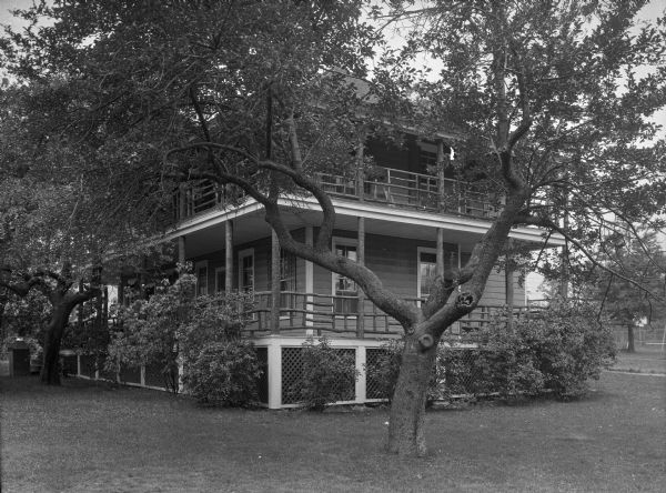 View across lawn toward Cottage U. Shrubs grow close to the porch, with tress in the lawn. A chair is on the upper veranda.