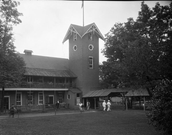 View across lawn toward the side of the Cottage and its corner tower. Part of the covered walkway is on the right. Women, men, and children are walking the grounds.