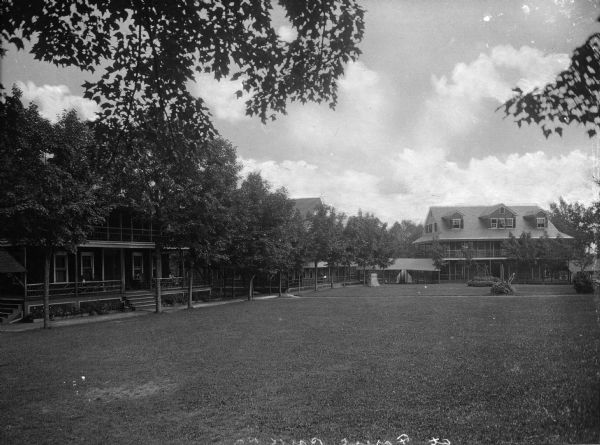 A view of the cottages, from the lawn. All the facades face inward onto this lawn lined with trees and shrubs. A woman sits on the porch on the cottage to the left side.