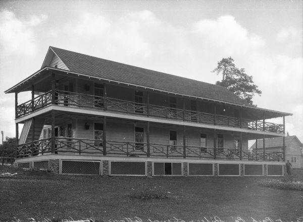 A view of the International Cottage, taken from the side. Chairs are on both porches, and a man stands to the right of the building near shrubs and flowers.