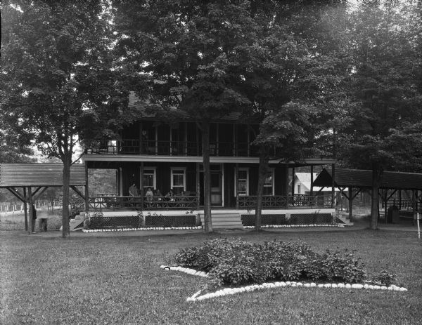 A view of the facade of Cottage T. Women and children sit on the porch.  The covered walkway is on both sides of the cottage. In front of the building is a planting of flowers with rocks forming an outline around it.