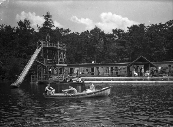 A view of people in row boats, posing for the camera. A man stands at the diving platform and slide. To the right, in a cordoned-off section, are swimmers. People also sit on the deck of the pavilion, which is covered, in both bathing suits and summer clothes. The background structure appears to be a series of changing rooms, with a clothesline in front.