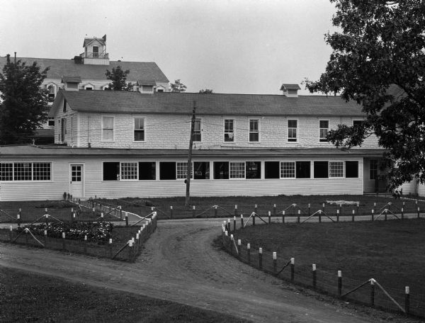 A view of the dance hall, with its many windows. On the second floor, a shirt drys in the window. The main house is in the background. Grassy areas and flower plantings border the dirt roads.