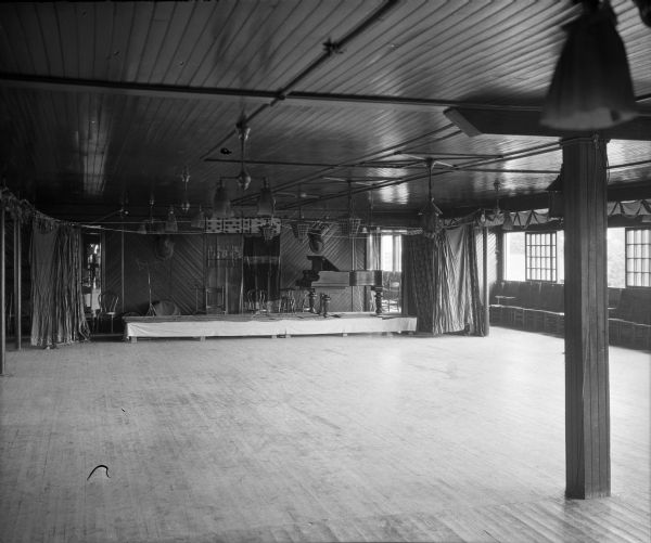 A view of the dance hall interior. At the back of the room is the stage, with curtains hung on either side. On the stage are stools and a piano. Large windows are along the right wall, with chairs in a row below. Electric lights line the ceiling.