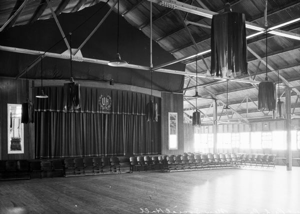 A view of the social hall and its stage. A special Unity House curtain is pulled across the front of the stage. Chairs are in rows lining the base of the stage. There are rafters and electric lights suspended from the ceiling.
