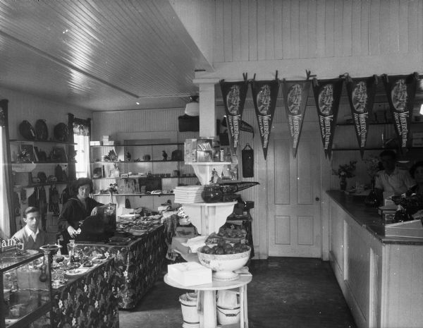 Slightly elevated view of the store's interior, with people posing behind the counters. Some of the items for sale are glassware, fruit, and souvenir pennants.