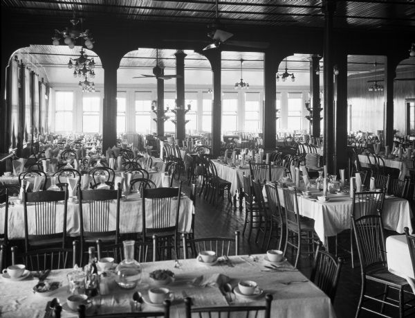 A view of the dining room with chairs around tables with place settings. The tables are arranged in rows, each set with silverware, cups and saucers, and water carafes. Fans and lighting fixtures hang from the ceiling. Brackets attached to the pillars have flower pots sitting on them.