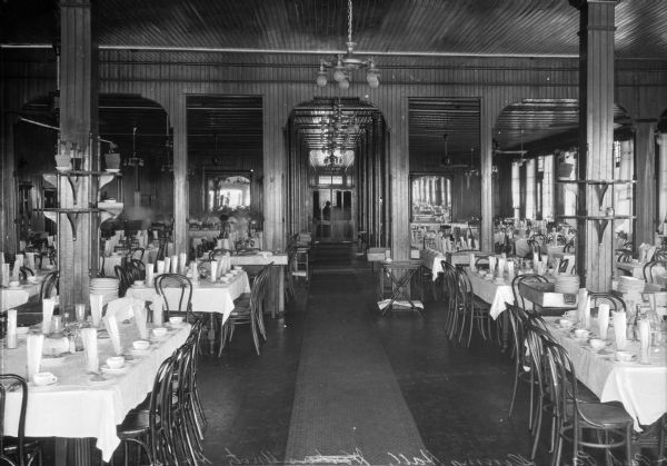 View down center of the dining room, looking toward the entrance door. The hall leading from the door to the dining room proper has a long carpet running its length. Brackets that are attached to pillars have flower pots sitting on them. The tables are set with silverware, glasses, water carafes, napkins, and cups and saucers. Fans and light fixtures line the ceiling.