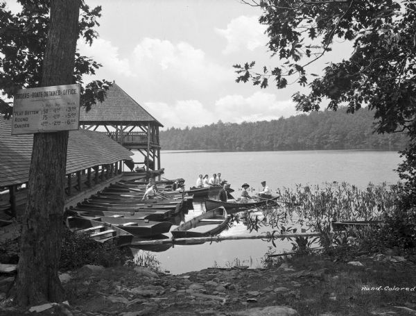 View toward shoreline with a covered pier and a pavilion on the end on the left. To the right of the pier are men and women in rowboats. A sign on a tree in the foreground on the left reads: "Orders for Boats Obtained at Office."
