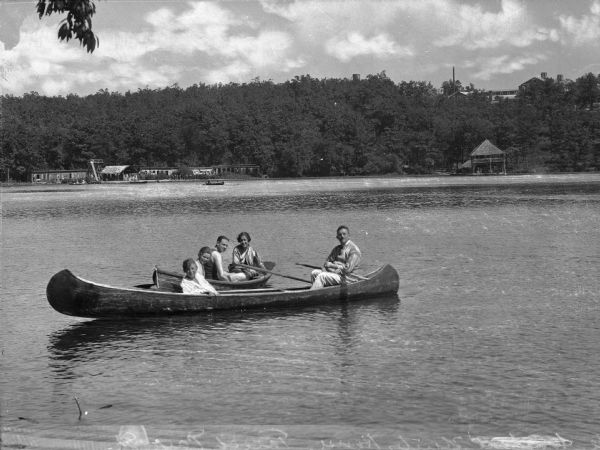 View across water toward women and men canoeing in two canoes. One canoe contains two women and a man; the other a woman and man. In the background on the shoreline on the left are changing rooms and a dock area with people. On the right is the boathouse.