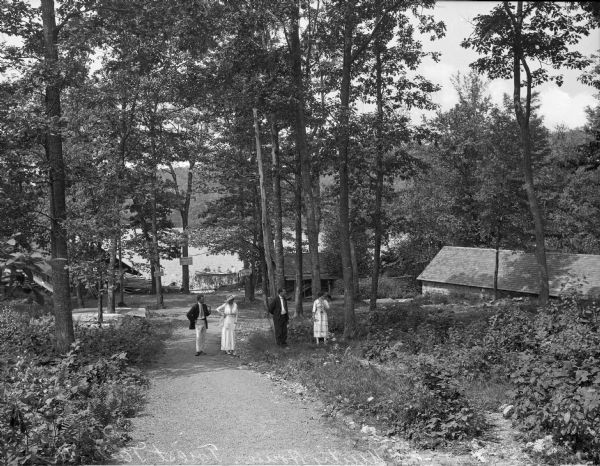 Elevated view of two couples walking on a trail. A lake is is the background, along with log cabins. A canoe with three people is in the background.