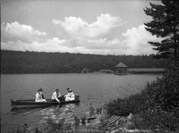 Two women and three men sitting in rowboat not far from the shore, posing for the camera. The dock and changing houses are in the background on the far shoreline.