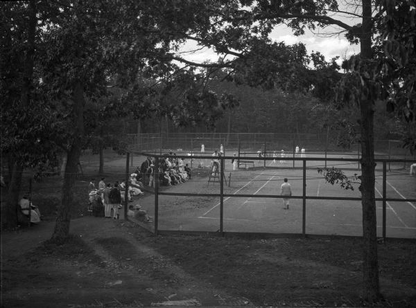 A view of the tennis courts set among trees. The court in the foreground has a game in progress, with men and women watching on the sidelines.