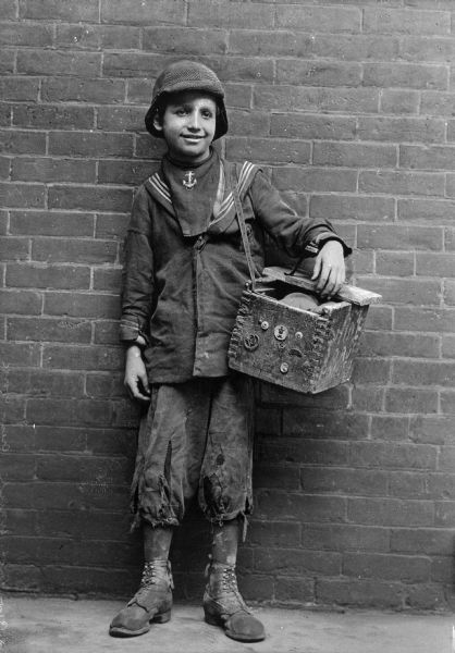 A portrait of a shoeshine boy, with a brick wall forming the backdrop. His clothes are tattered, and his work box is hanging from a strap on his left shoulder.