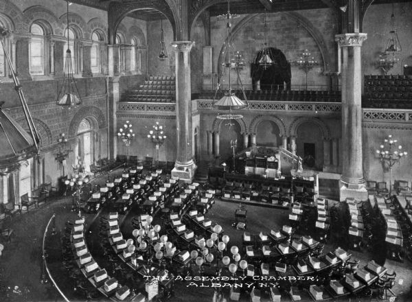 Elevated view of the Assembly Chamber interior. The desks and speaker's podium are below in the center. Large light fixtures in the ceiling and on the floor are abundant. Printed at the bottom of the image: "The Assembly Chamber, Albany, N.Y."