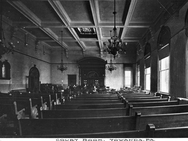 A view of the interior, taken from the audience benches, looking toward the judge's podium.  Five men pose from the front of the court room, on both sides of the podium.  A stained glass skylight is visible in one of the coffers.  Printed below the image: "Court Room, Towanda, PA."