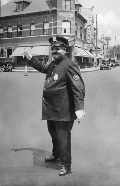 A portrait of a police officer directing traffic with a pleasant look on his face and one hand in the air. A building on the corner behind the man reads "5 Corners Hotel." Automobiles are parked along the curbs.