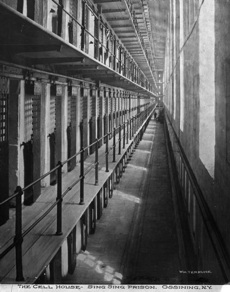 A interior view of a cell block. Cells line the left side, with five levels visible. Long rectangular windows line the wall to the right.  Two men, presumably guards, are visible on the first floor, by the stairs. Caption reads: "The Cell House - Sing Sing Prison, Ossining, NY."