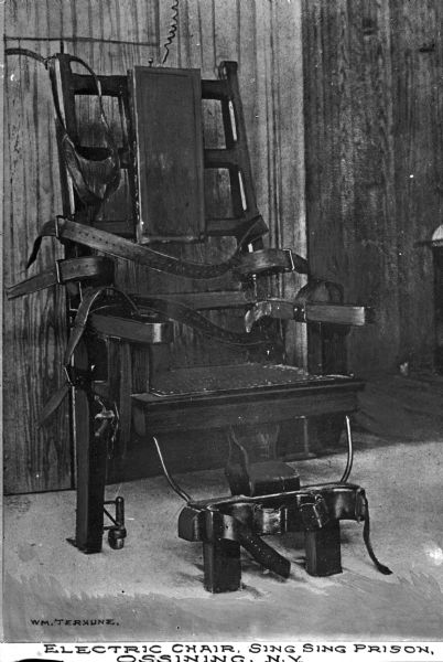 View of an electric chair with straps in a room with wooden walls and  a concrete floor. Caption reads: "Electric Chair, Sing Sing Prison, Ossining, N.Y."