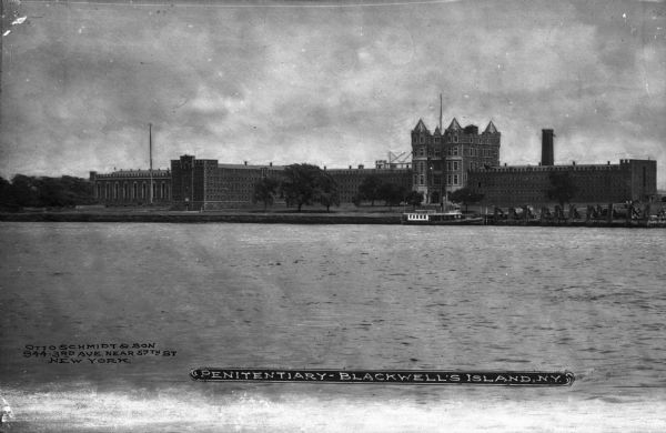 View across river toward the exterior of the prison, with the central tower and wings. A boat sits at the dock on the far shoreline, and a large smokestack rises behind the building. Caption reads: "Penitentiary-Blackwell's Island, N.Y."