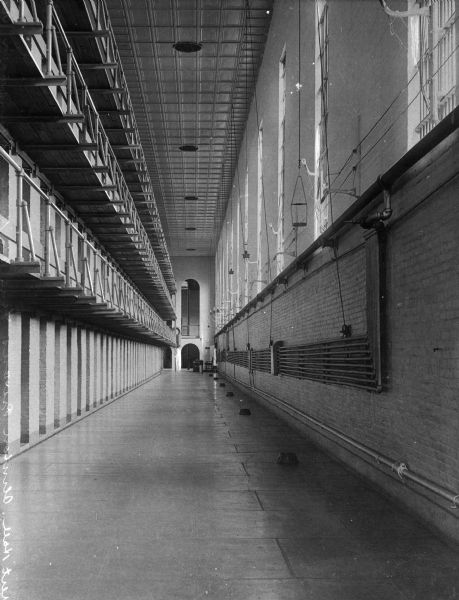 A view at ground floor of the cell block, with cells on the left and windows on the right outer wall; a series of pipes also run along this wall. An arched door and window are at the other end of the corridor in the background.