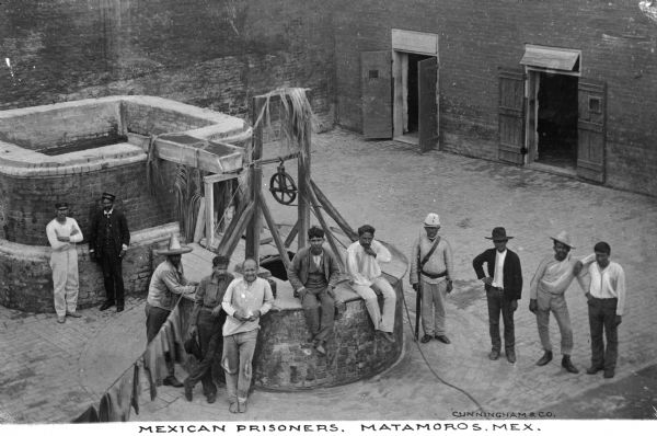 Elevated view of prisoners posing by the washing well, in a courtyard. Some of the men wear hats. Wooden doors of a building are on the right. Caption reads: "Mexican Prisoners, Matamoros, Mex."