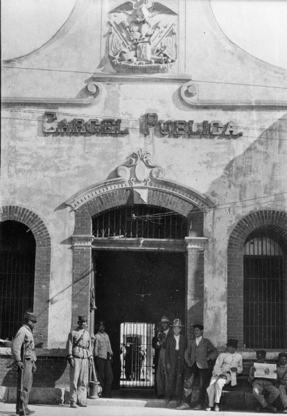A view of the Juarez, Mexico city jail. Officers and other men stand in front of the structure, made of adobe. Brick arches trim the windows and doorway. Other stone details are above the doorway and in the pediment area. The wall has a sign that reads: "Carcel Publica."