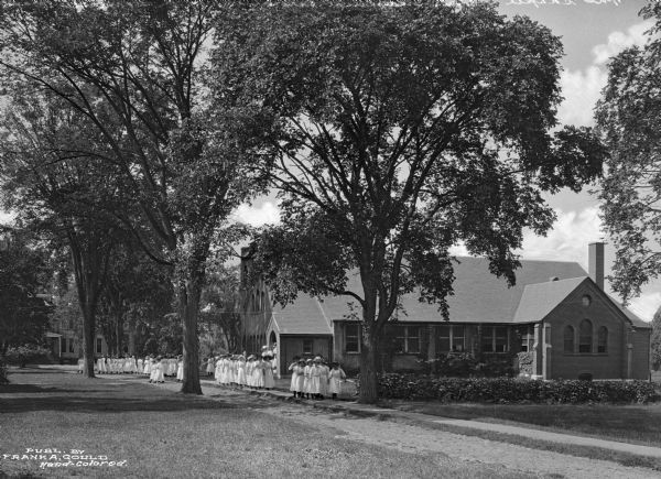 A view of the chapel and dining hall of the school, with another structure partially hidden by trees in the background, to the left.  Females in white uniforms stand on the path in front the chapel.