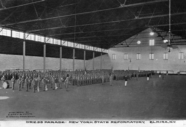A view of a dress parade, with the men wearing military-like uniforms.  They men are lined up in orderly rows in a field house. Caption reads: "Dress Parade — New York State Reformatory, Elmira, N.Y."