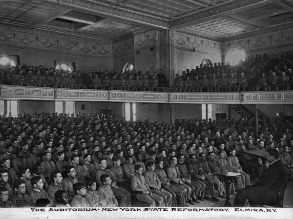 View from stage of the auditorium. Male inmates fill the seats, and a man sits on the stage facing them. Caption reads: "The Auditorium - New York State Reformatory, Elmira, N.Y."