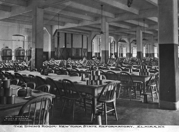 A view of the dining room. There is a utensil and plate at each seat, with cups and bowls grouped at the head of the table, with a large serving utensil. Caption reads: "The Dining Room-New York State Reformatory, Elmira, N.Y."