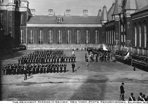 Elevated view of the regiment, lined up in a courtyard. Men with firearms face the viewer, while other men stand to the right side. The band stands at the right, in front of the men. The walls of the reformatory are prominently seen on the right and in the background. Caption reads: "The Regiment Passing in Review-New York State Reformatory, Elmira, N.Y."