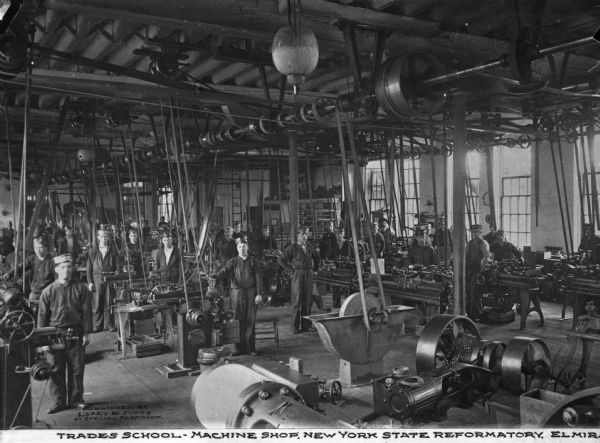 A view of the trade school machine shop, with a man standing by each machine. Light streams in from the windows on both sides of the shop. Caption reads: "Trade School - Machine Shop, New York State Reformatory, Elmira, N.Y."
