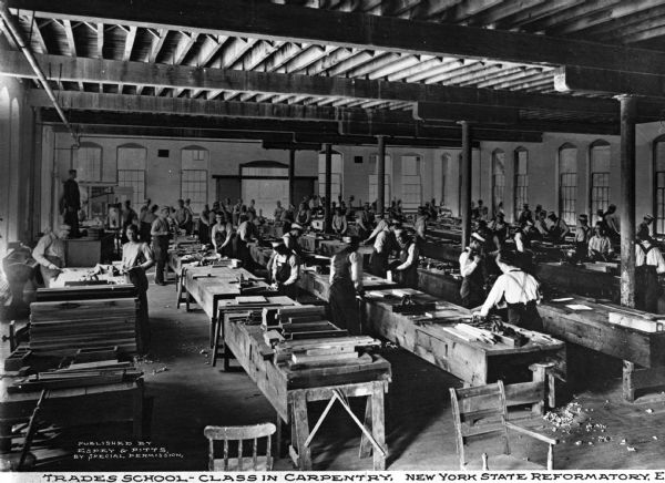 A view of  a trade school carpentry class. Men are in the process of working at their benches. Light comes in through the windows to the left. Caption reads: "Trades School - Class in Carpentry, New York State Reformatory, Elmira, N.Y."