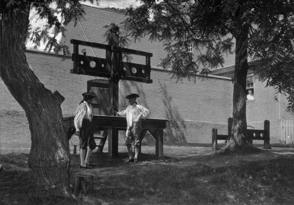 A view of two costumed interpreters standing by a pillory in Colonial Williamsburg. A man poses in a window of the building to the right.