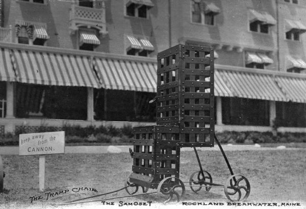 A view of a chair on wheels with a hotel in the background. A sign on the lawn reads: "Keep away from the Cannon." Printed near the bottom left corner: "The Tramp Chair." In the middle: "The Samoset," and text at bottom right corner: "Rockland Breakwater, Maine."