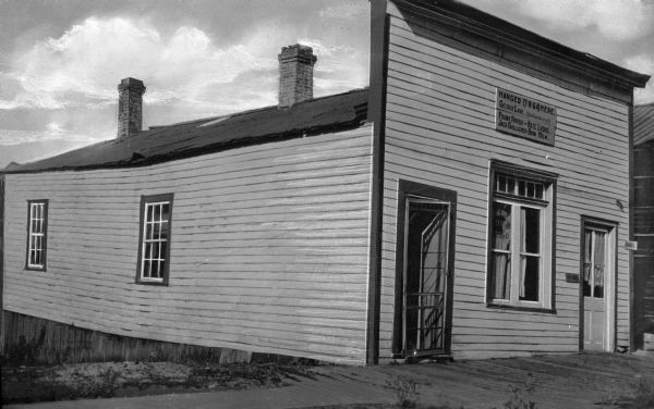 View toward the front and left side of a clapboard house, which has a window in the middle of the facade and a door flanking each side. A sign above the window reads: "Hanged (1864) Here: George Lane (Club Foot George). Frank Parrish — Haze Lyons. Jack Gallagher — Boon Helm."