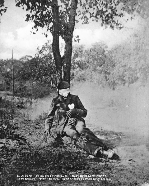 View of a man executed by firing squad at the base of a tree. His face is covered with a cloth, and he has a small heart bull's eye on his chest.