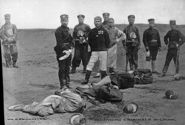 A view of four beheaded men lying on the ground with a group of officers standing behind, posing for the camera. A man to the left in the central group holds a knife out, with a rag over it. Caption reads: "Beheading Criminals in China."