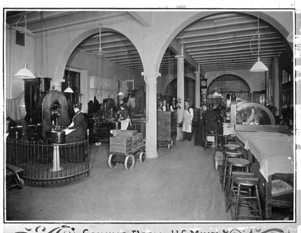 A view of the Coining Room, with men and women working at various machines. Large pallets of boxes, on wheels, sit in the central aisle, on the left. Caption reads: "Coining Room — US Mint."