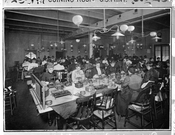 A view of the Coining Room, where women sit at long tables, weighing out metal.  Each woman has a balance and other tools at her station.  A male supervisor stands near a column in the back of the image to observe.  Printed above the image: "Coining Room-U.S. Mint."