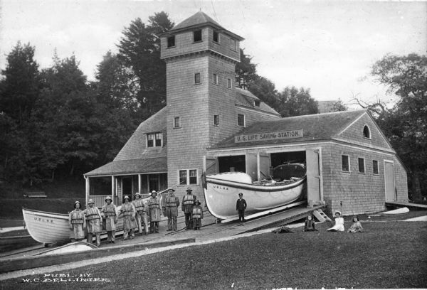 A view of the boathouse and boats used by the station men, who pose in their gear. Three girls and one boy pose off to the right of the scene; the girls sit on the grass while the boy stands next to another boat, sitting in the open doorway of the boathouse.