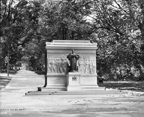 A view of the monument, which contains, in low relief, men as civilians and as soldiers. In front of this is a sculpture of an angel with a sword in her hand. Behind the monument is a tree-lined path.