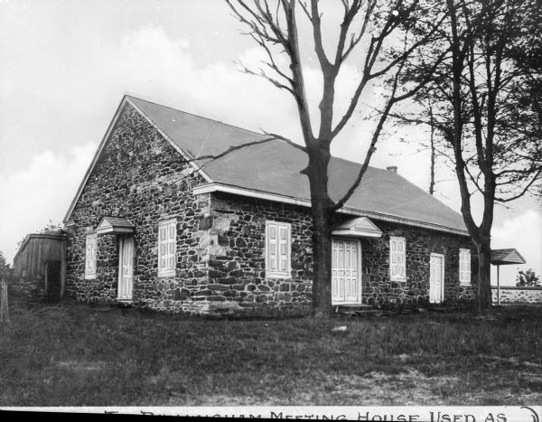 View toward front and left side of the meeting house constructed of field stone, with white shutters and doors on both sides. A wooden addition is on the back, to the left, as there is a stone wall in the background on the right. Caption reads, in part: "[The Birmingham] Meeting House Used As [...]"