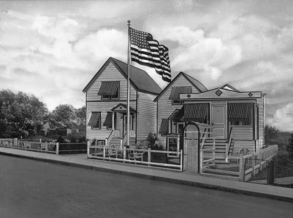 View across road toward the church, which consists of two small buildings. The windows have awnings, and the yard is fenced. To the left is another building with awnings on the windows and a fenced area. A large flag on a flagpole is in the yard near the sidewalk.