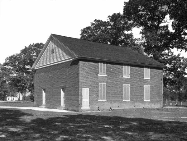 View across lawn toward the front and right side of the brick church. The front has two doors, and the side has six shuttered windows.