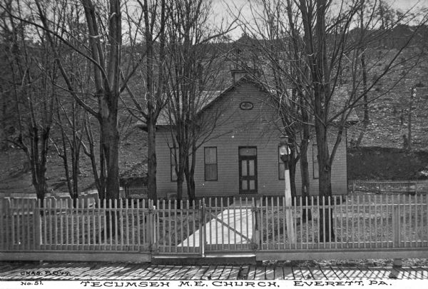 View over sidewalk and picket fence toward the church exterior surrounded by trees. The path leads from the sidewalk to the door whcih is flanked by two windows on each side. Caption reads: "Tecumseh M. E. Church, Everett, PA."